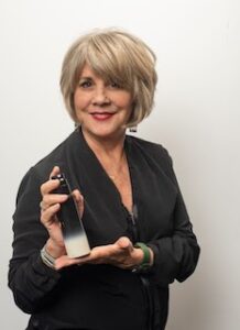 A person with short hair and a black jacket holds a spray bottle, smiling at the camera.