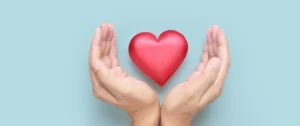 Two hands are positioned around a red heart-shaped object against a light blue background.