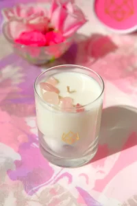 A white candle in a glass jar with pink crystals embedded in it, placed on a pink and white surface. In the background, a bowl of pink rose petals is visible.