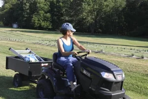 A person wearing a blue tank top and cap is sitting on a black lawnmower with a trailer attached, in a field with rows of plants and trees in the background.