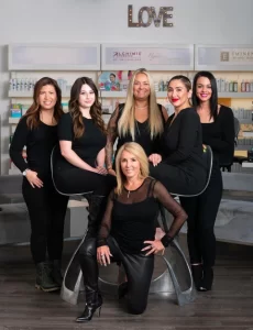 A group of six women in black attire posing inside a beauty salon, with shelves of beauty products and a sign that reads "LOVE" in the background.