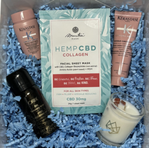 A gift box containing a Hemp CBD collagen facial sheet mask, two Kérastase products, a bottle of Oribe product, and a white candle, all nestled in blue shredded paper.
